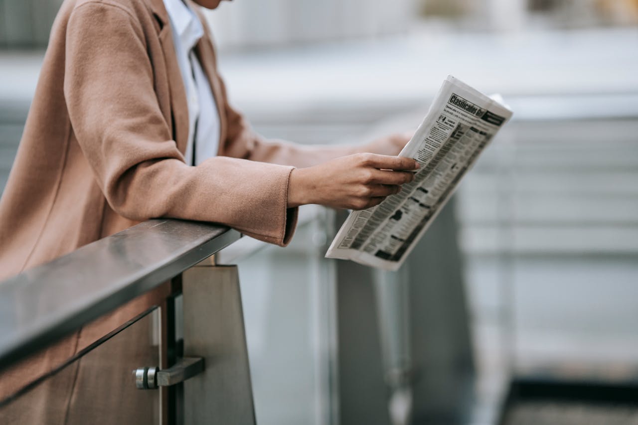 A well-dressed woman leans on a railing, engrossed in a newspaper outdoors.